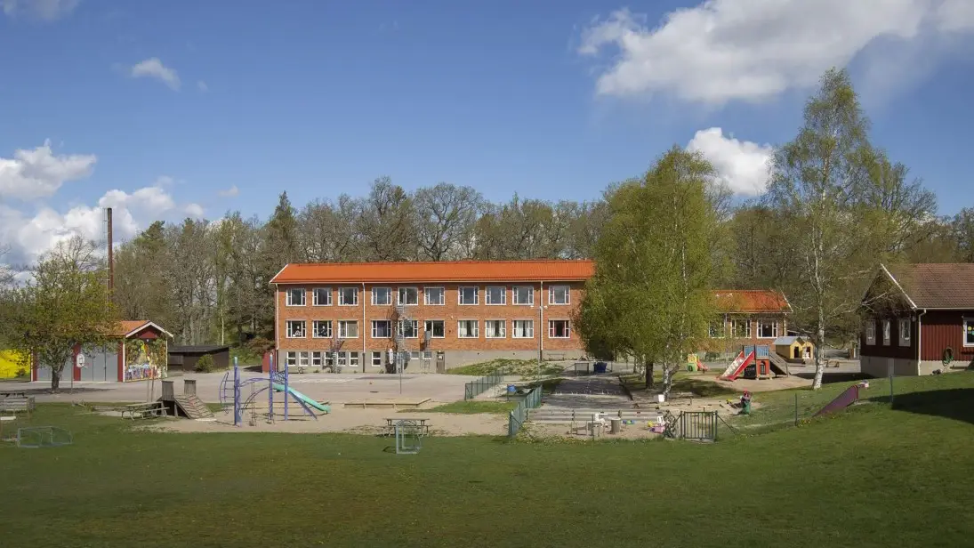En tegelbyggnad i tre våningar. I förgrunden en skolgård med lekplats och gräsmatta. I bakgrunden en träddunge.