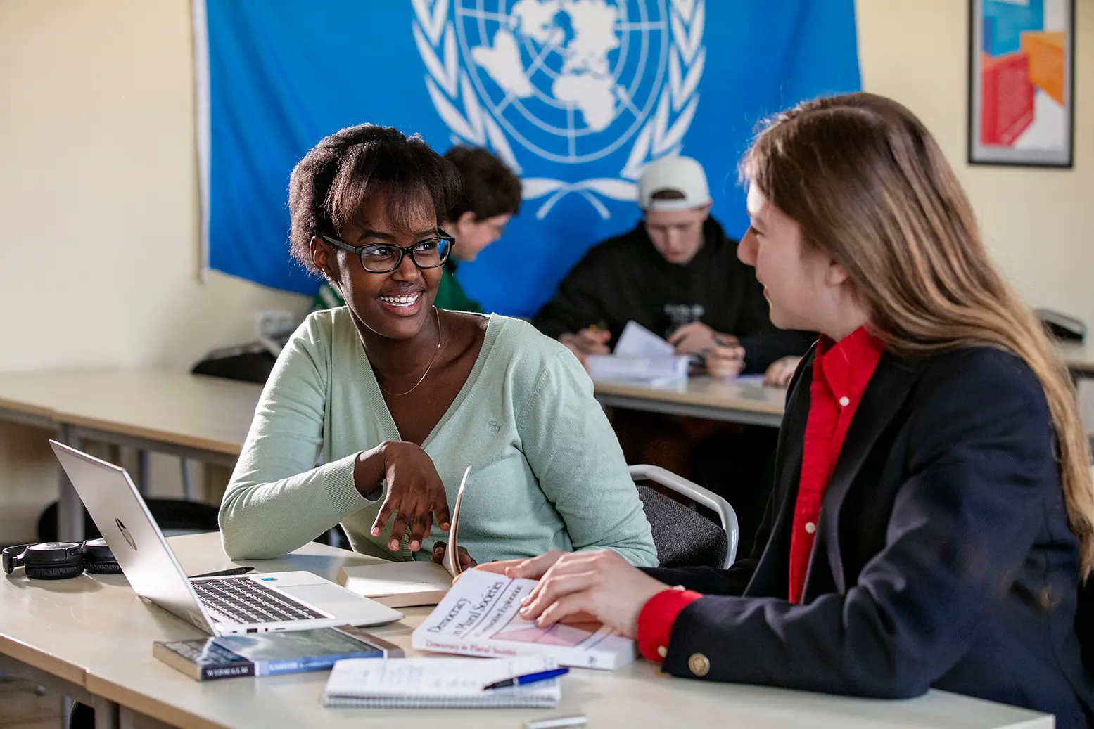 Två elever i ett klassrum med böcker och dator framför sig. På väggen bakom hänger FN-flaggan
