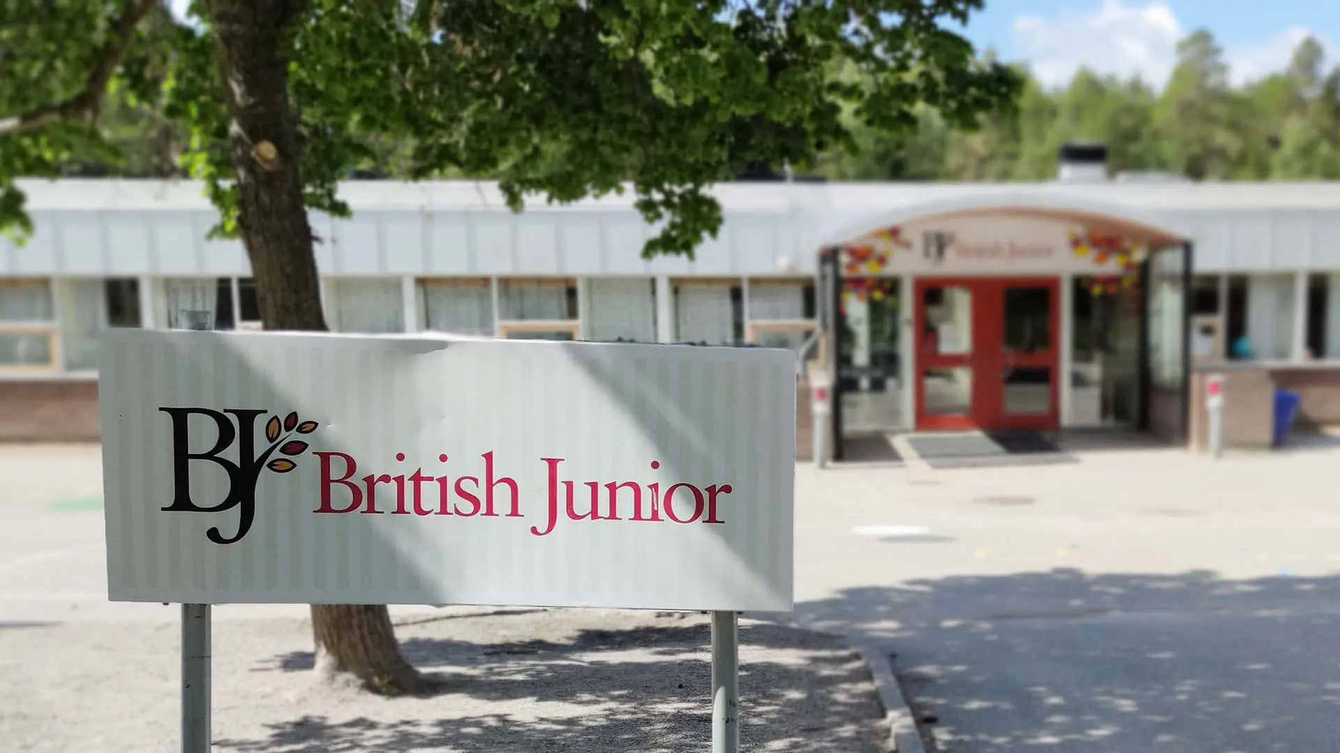 Skolbyggnad i en plan i bakgrunden. Skylt med British Juniors logotyp i förgrunden.