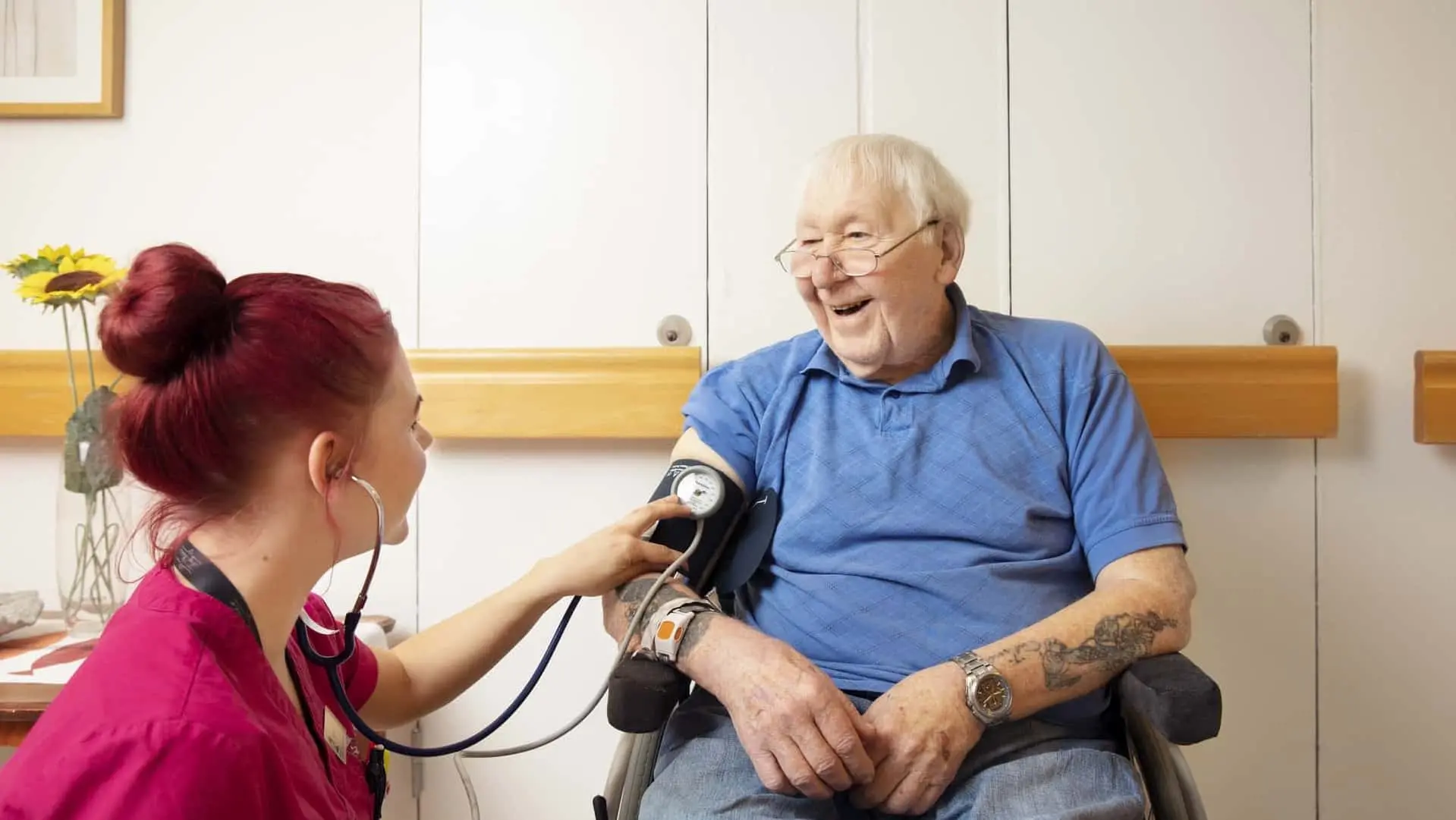 Kvinna med stetoskop använder stetoskop på armen på en glad man i rullstol.
