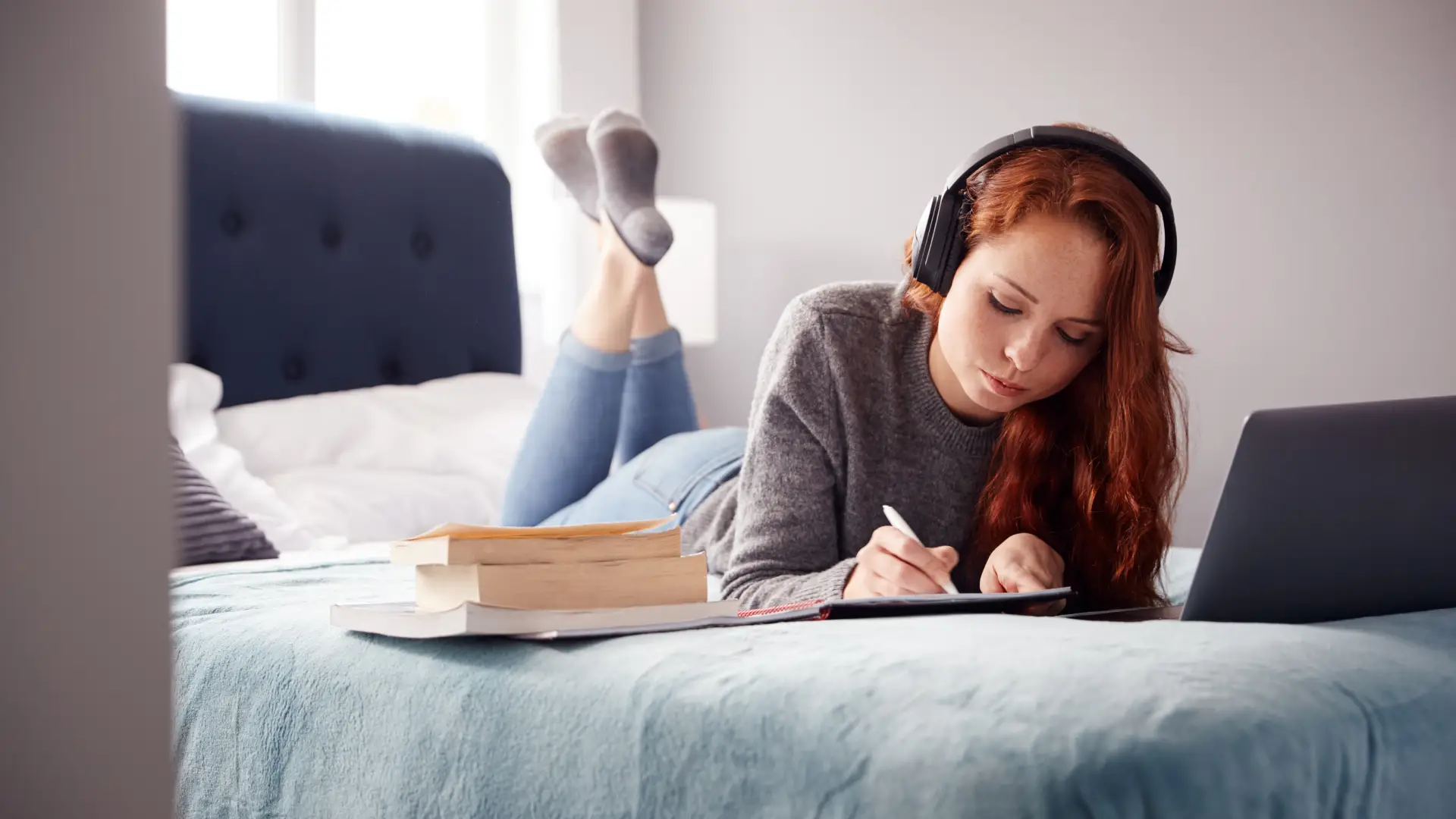 En kvinnlig student ligger på en säng och skriver. Framför sig har hon en dator och bredvid sig flera böcker.